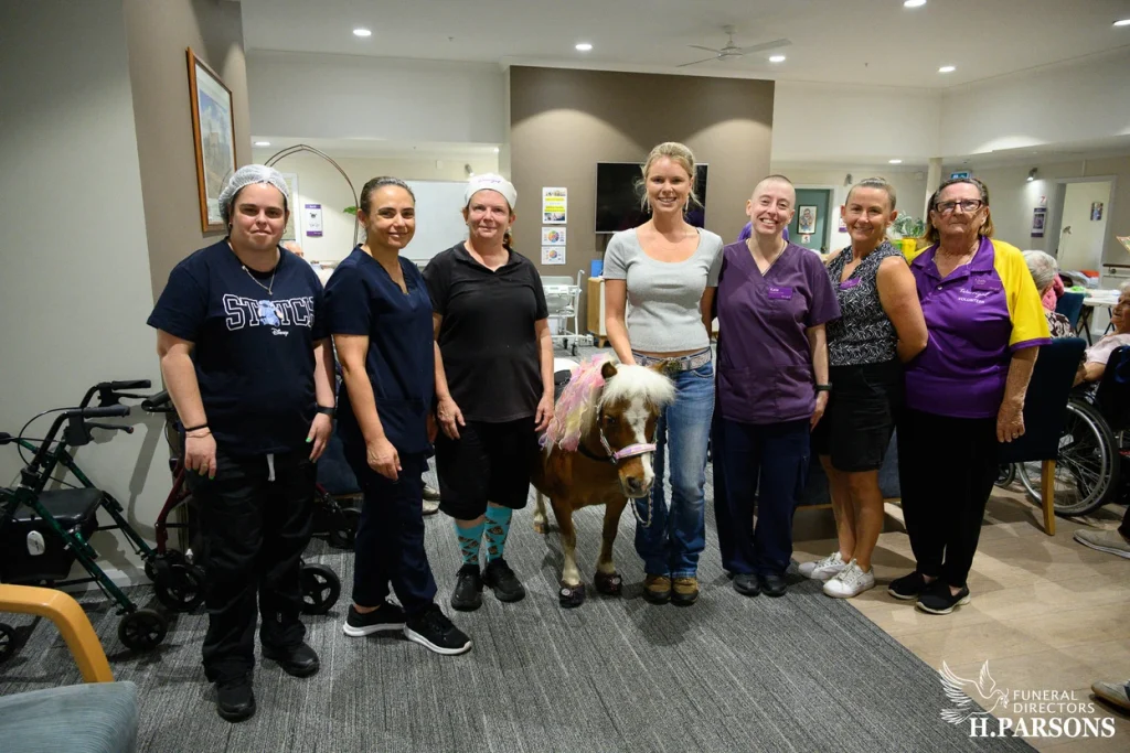 A group of seven adults stand indoors, smiling at the camera with a small pony from Therapy Horses Illawarra in the centre. The setting appears to be a care facility or nursing home, with medical equipment and furniture visible in the background. by H Parsons Funeral Directors