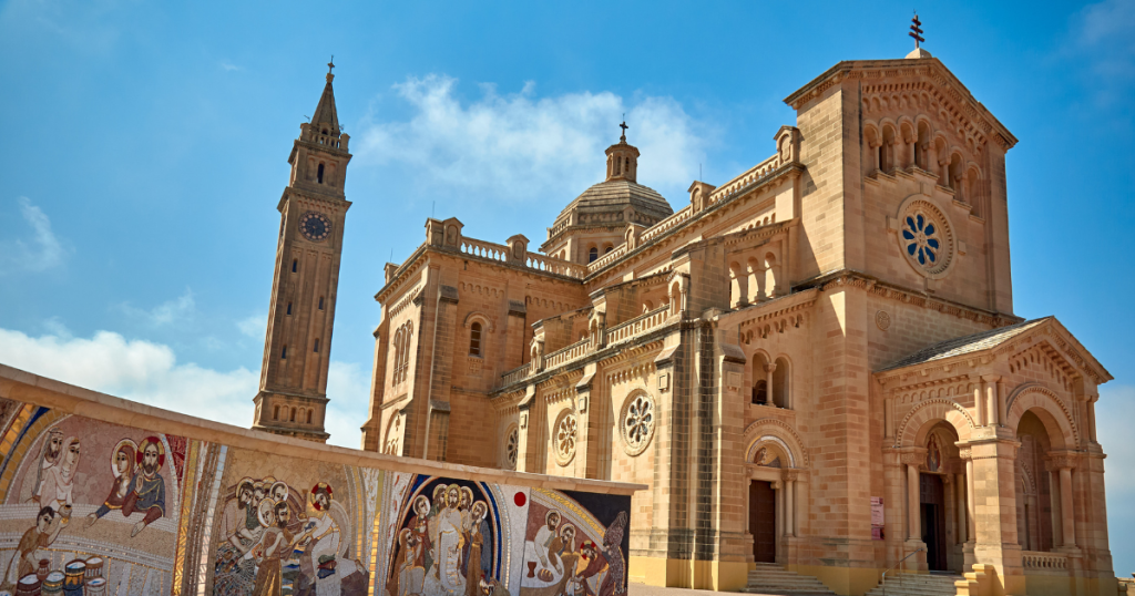 A large sandstone church with a central dome and tall clock tower stands under a blue sky; a colourful mosaic depicting religious scenes in the foreground reflects Greek funeral traditions. by H Parsons Funeral Directors