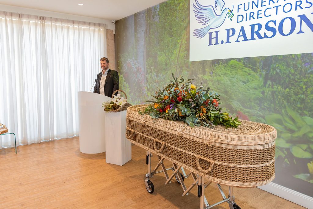 A man stands at a lectern in a bright room with a coffin adorned with flowers in the foreground and a large "H. Parsons Funeral Directors" sign on the wall behind him, reflecting evolving funeral trends Australia now embraces. by H Parsons Funeral Directors