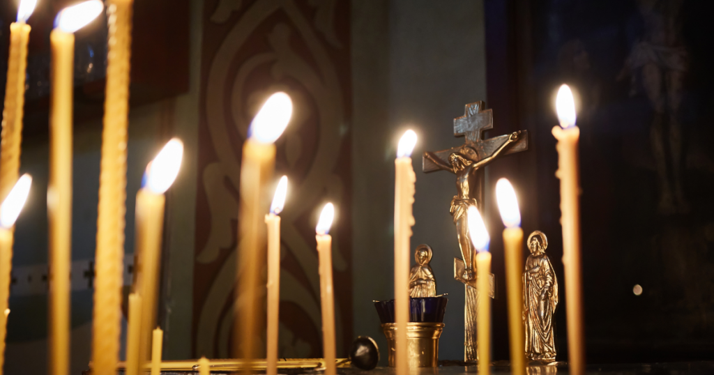 Lit candles in front of gold religious icons, including a crucifix and small saint statues, set against a dimly lit background with decorative patterns—an atmosphere often seen during Orthodox funeral traditions. by H Parsons Funeral Directors