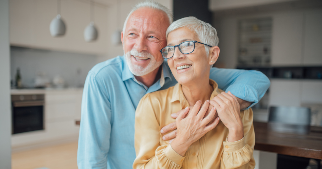 An older couple smiles and embraces in a bright, modern kitchen. The man, with grey hair and a beard, stands behind the woman, both appearing happy and relaxed—showing the peace of mind that comes from pre-planning important moments together. by H Parsons Funeral Directors