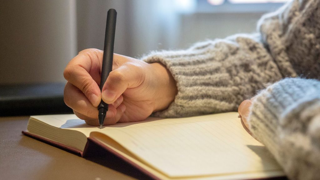 A person wearing a grey knitted jumper is writing in a lined notebook with a black pen, perhaps reflecting on how long a eulogy should be as they compose their thoughts. The focus is on their hand and the notebook, with a soft, blurred background. by H Parsons Funeral Directors
