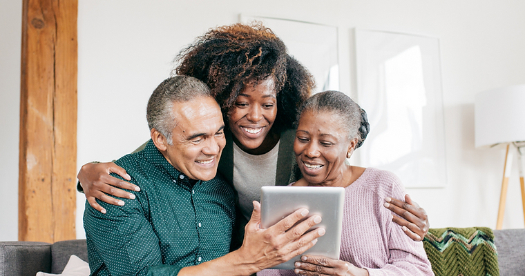 Three people sit on a sofa smiling and looking at a tablet together. The two older adults hold the tablet while a younger woman stands behind them, perhaps exploring ideas for celebration of life on the screen. by H Parsons Funeral Directors