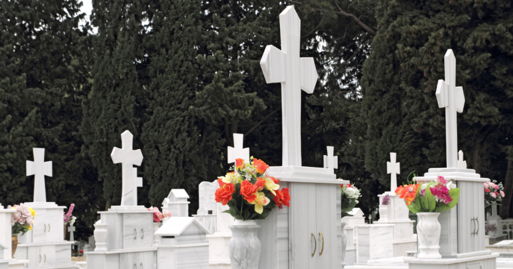 Rows of white marble graves with large crosses, each adorned with colourful flower arrangements in vases, reflect Greek funeral traditions and the sombre beauty typical of an Orthodox funeral, set against tall, dark green trees in a peaceful cemetery. by H Parsons Funeral Directors