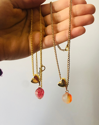 A hand holds two gold chain necklaces, each with a heart charm and a teardrop-shaped pendant—one pink and the other white with orange specks—making thoughtful keepsakes or unique ideas for celebration of life moments. by H Parsons Funeral Directors