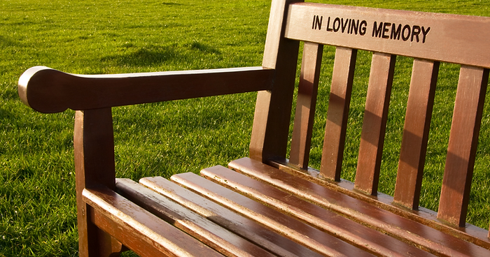 A wooden bench in sunlight on a grassy lawn, engraved with "IN LOVING MEMORY" on the backrest—an inspiring memorial idea for those seeking unique celebration of life ideas. by H Parsons Funeral Directors