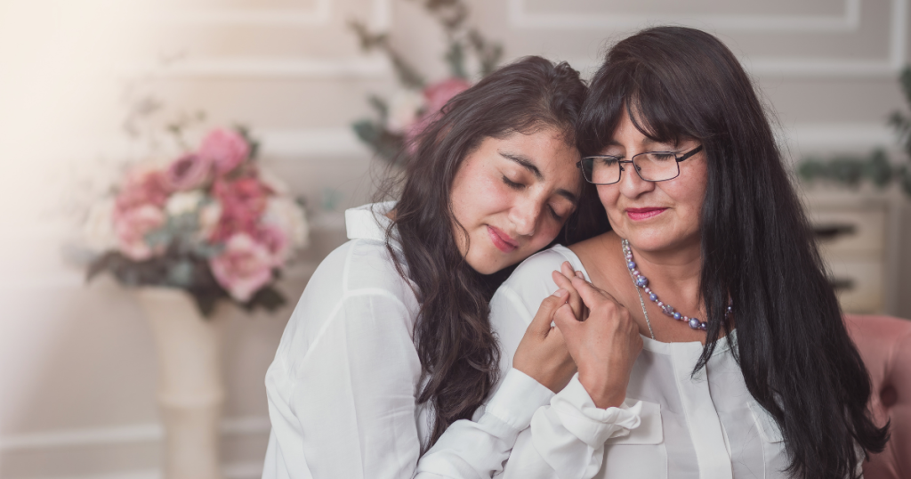 A young woman, grieving on Mother’s Day, rests her head on an older woman’s shoulder while holding her hand. Both wear white tops and soft smiles as they sit closely together, surrounded by gentle floral decorations. by H Parsons Funeral Directors