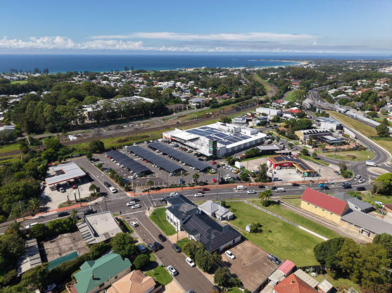 Aerial view of a coastal suburban area featuring a shopping centre with a large car park, surrounding roads, houses, greenery, and the sea visible in the background under a blue sky. by H Parsons Funeral Directors
