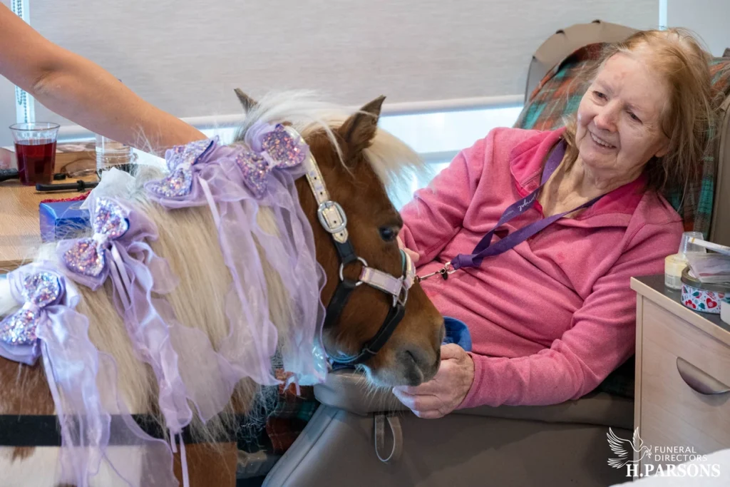 An elderly woman sits and smiles while stroking a small horse dressed in a purple costume with bows. The therapy horse, part of Therapy Horses Illawarra, is inside the room, bringing joy as shown by the woman's happy and engaged expression. H. Parsons logo visible. by H Parsons Funeral Directors