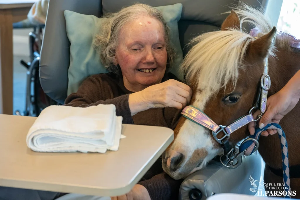 An elderly woman smiles while petting a small therapy Illawarra horse beside her chair. A folded towel is on a tray in front of her, and a person holds the horse’s reins as its head rests gently on her lap. by H Parsons Funeral Directors