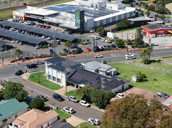 Aerial view of a street crossroads with solar-panelled buildings, a supermarket with a large car park, and several cars driving and parked around the area. Green lawns and trees are visible throughout the scene. by H Parsons Funeral Directors