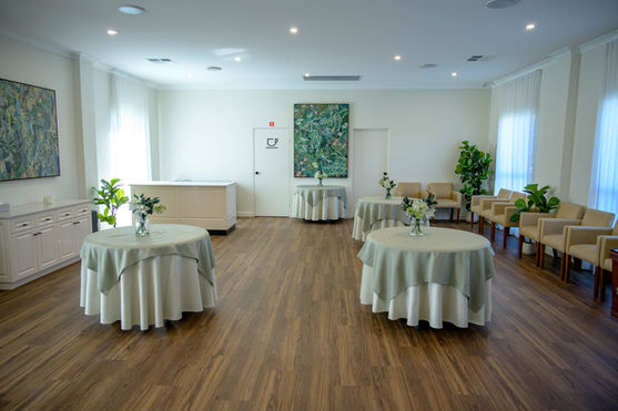 A bright, elegant room with wooden floors, three round tables covered in light tablecloths, chairs arranged along the walls, potted plants, large paintings, and soft natural light from tall windows. by H Parsons Funeral Directors