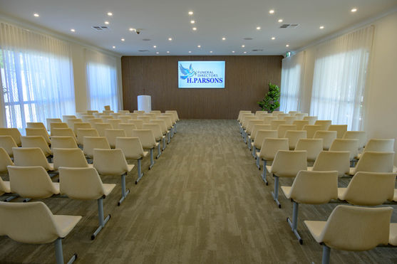 A modern, well-lit room with rows of beige chairs facing a lectern and a large screen displaying "H. Parsons." The space has carpeted floors, cream-coloured walls, and large windows with sheer curtains. by H Parsons Funeral Directors