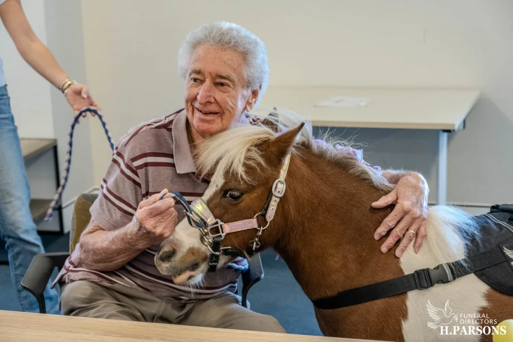 An elderly man with grey hair, wearing a striped polo shirt, smiles while stroking a small brown and white therapy horse from Therapy Horses Illawarra indoors. A person holds the pony’s lead rope beside him. by H Parsons Funeral Directors