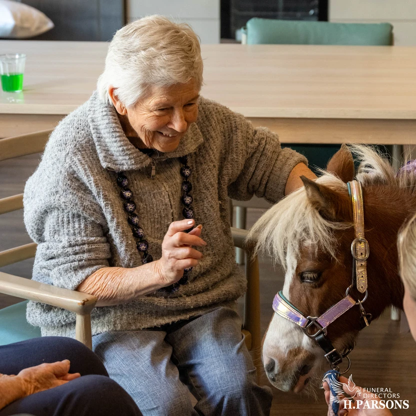 An elderly woman with white hair smiles and gently strokes a small brown and white horse from Therapy Horses Illawarra indoors. Another person sits nearby, with a drink on the table behind them. by H Parsons Funeral Directors