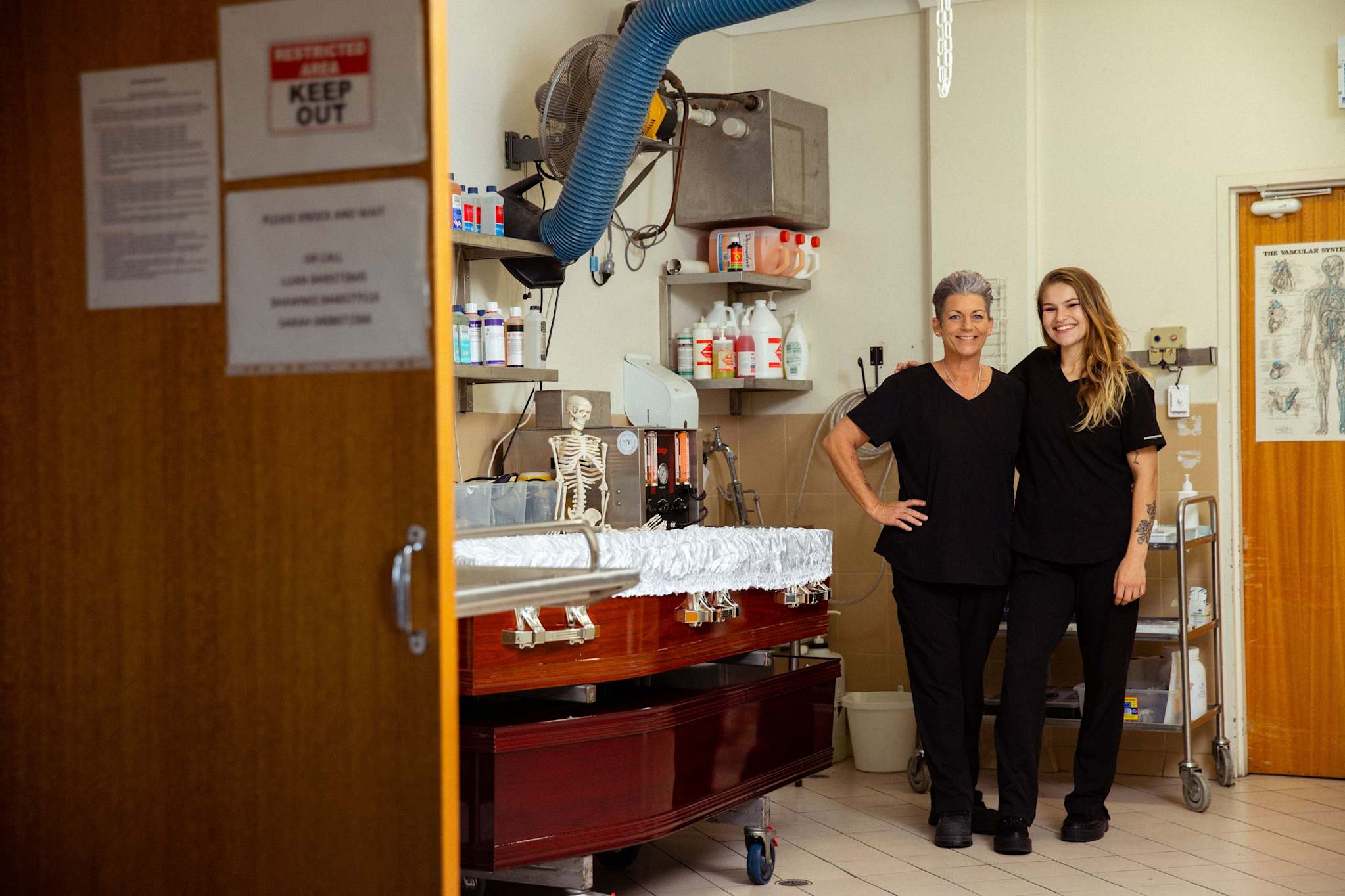Two women in black uniforms stand smiling beside a coffin in a mortuary preparation room. Supplies and equipment are visible around them, and a "Keep Out" sign is posted on the open door in the foreground. by H Parsons Funeral Directors