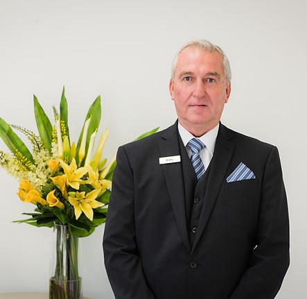 A man in a dark suit with a striped tie and name tag stands beside a table with a vase of yellow lilies and greenery, against a plain white background. by H Parsons Funeral Directors