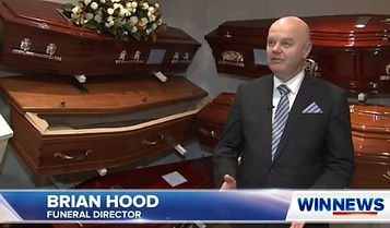 A man in a suit stands in front of several wooden coffins, speaking. Text overlay reads "Brian Hood, Funeral Director" and "WIN News" on the bottom of the image. by H Parsons Funeral Directors