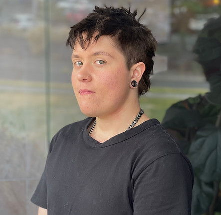 A person with short, dark, tousled hair and light skin stands indoors, wearing a black t-shirt, chain necklace, and black ear plugs. They look slightly to the side with a neutral expression. by H Parsons Funeral Directors