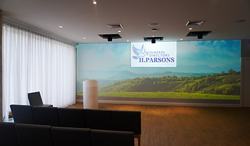 A modern funeral service room with rows of black chairs facing a screen displaying "FUNERAL DIRECTORS H. PARSONS" above an image of a peaceful green landscape and hills, with natural light streaming through white curtains. by H Parsons Funeral Directors