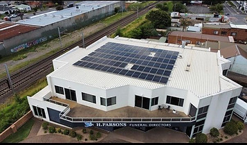 Aerial view of a white building with solar panels on the roof, next to a railway line. The sign on the building reads "H. Parsons Funeral Directors." Surrounding areas include roads, grass, and industrial buildings. by H Parsons Funeral Directors