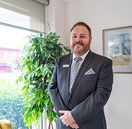 A man in a suit and tie stands indoors near a window and a potted plant, smiling at the camera. He wears a name badge and has a pocket square in his jacket. A framed picture hangs on the wall behind him. by H Parsons Funeral Directors