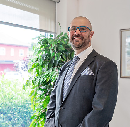 A man wearing a dark suit, striped tie, and blue glasses stands smiling in a bright office next to a large green plant and a window with natural light coming in. by H Parsons Funeral Directors
