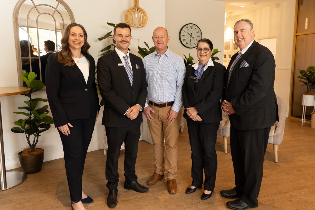 Five professionally dressed adults stand smiling in a well-lit, modern office space with plants, a wall mirror, and a clock in the background. Four wear suits, one is in a light blue shirt and chinos. by H Parsons Funeral Directors