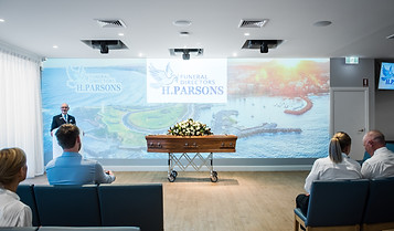 A funeral service with a coffin and flower arrangement at the front, a speaker at a lectern, and attendees seated. The background displays an image of a coastal landscape and a sign reading "H. Parsons Funeral Directors. by H Parsons Funeral Directors