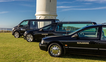 Three black hearses with "H. PARSONS" written on the side are parked on grass near a white columned structure, with the sea visible in the background. by H Parsons Funeral Directors