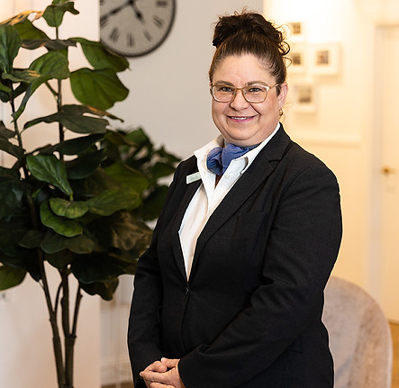 A woman wearing glasses and a black suit with a blue scarf stands smiling indoors beside a large leafy plant. A clock and framed pictures are visible on the wall in the background. by H Parsons Funeral Directors