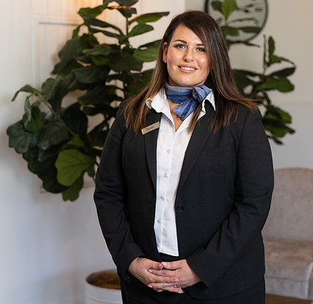 A woman in a dark suit and blue neck scarf stands indoors, smiling at the camera, with a large green plant and light-coloured furniture in the background. by H Parsons Funeral Directors