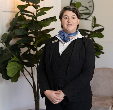 A person wearing a black suit, white shirt, and blue scarf stands indoors in front of a large leafy plant, smiling at the camera. A clock and chair are visible in the background. by H Parsons Funeral Directors