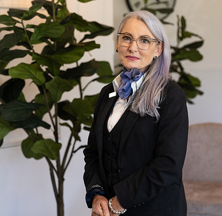 A woman with long grey hair, wearing glasses, a black suit, and a blue scarf stands indoors near green plants, looking at the camera with a slight smile. by H Parsons Funeral Directors