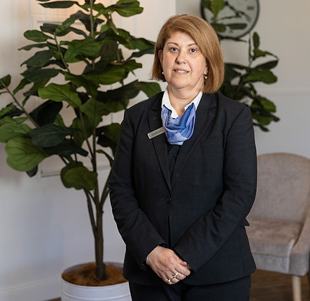 A woman in a black suit stands indoors in front of large potted plants. She has short light brown hair and wears a blue scarf and name badge, posing with her hands folded. The setting appears to be an office or reception area. by H Parsons Funeral Directors