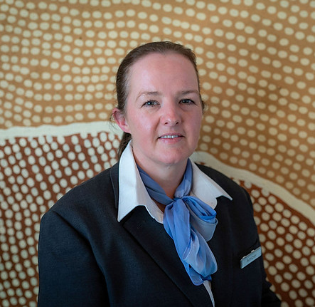 A woman in a dark blazer, white shirt, and blue scarf stands in front of a tan background with white dots and lines. She has light skin, brown hair pulled back, and is looking at the camera, smiling slightly. by H Parsons Funeral Directors