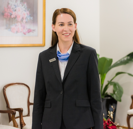 A woman in a black blazer and blue scarf stands indoors, smiling. Behind her are white chairs, a plant, and a floral painting on the wall. by H Parsons Funeral Directors