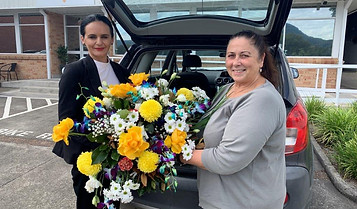 Two women stand by an open car boot, smiling and holding a large, colourful bouquet of flowers with yellow, white, and purple blooms, outside a building on a cloudy day. by H Parsons Funeral Directors