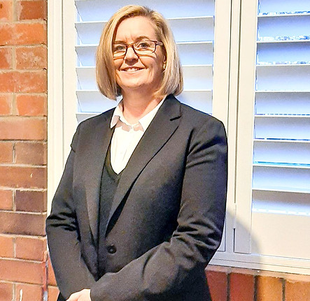 A woman with blonde hair and glasses, wearing a dark blazer over a white shirt, stands smiling in front of a window with white shutters and a brick wall. by H Parsons Funeral Directors