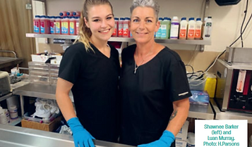 Two women wearing black shirts and blue gloves stand side by side in a kitchen or food preparation area, smiling at the camera. Shelves with bottles and supplies are visible in the background. by H Parsons Funeral Directors