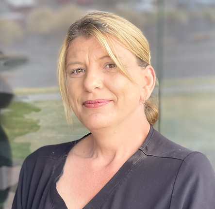 A woman with blonde hair tied back and wearing a black top smiles softly at the camera in front of a blurry outdoor background. by H Parsons Funeral Directors