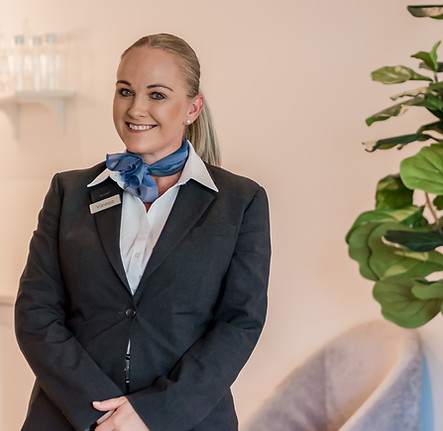 A smiling woman in a dark blazer, white shirt, and blue neck scarf stands indoors near a green leafy plant, with light-coloured walls and decor in the background. by H Parsons Funeral Directors