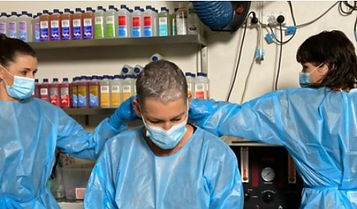Three people in blue protective gowns and face masks are in a laboratory or salon; one person sits with a shaven head while the other two stand on either side, working with their hands near the seated person's head. Bottles line shelves in the background. by H Parsons Funeral Directors