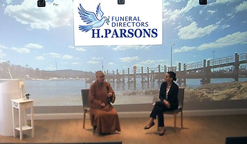 Two people sit on stage under a sign for "H. Parsons Funeral Directors." One is in a brown robe, the other in a suit. Behind them is a large scenic backdrop of a pier, water, and blue sky with clouds. by H Parsons Funeral Directors