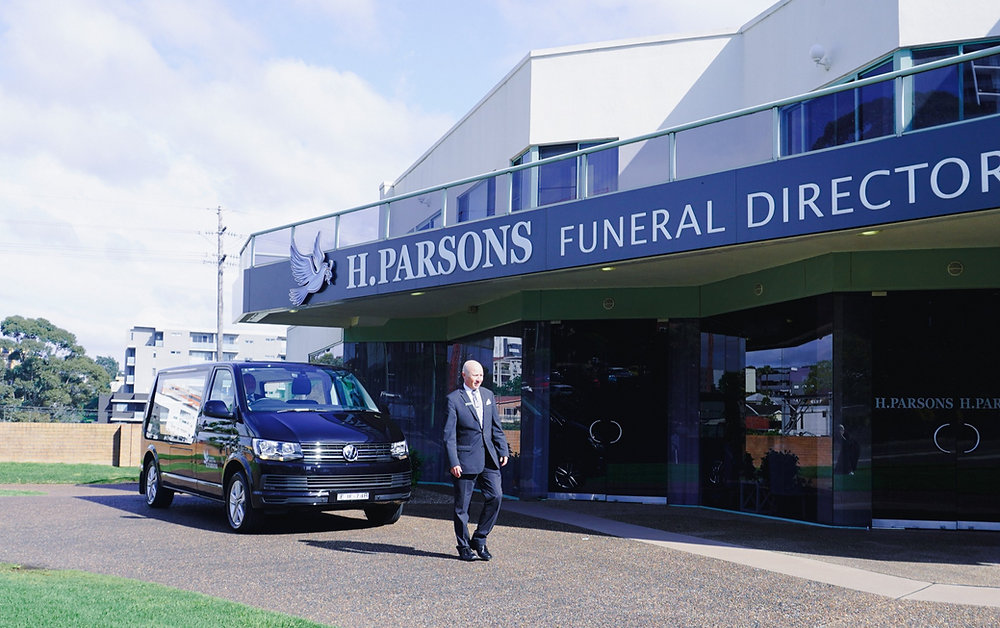 A man in a suit walks in front of a black van parked outside H. Parsons Funeral Directors, a modern building with large windows and a prominent sign offering funeral assistance. The sky is partly cloudy. by H Parsons Funeral Directors