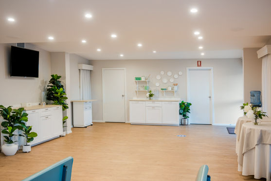 A bright, spacious room with wooden floors, white cupboards, potted plants, a wall-mounted TV, shelves with decor, and a water dispenser. The space appears clean and modern, lit by ceiling spotlights. by H Parsons Funeral Directors