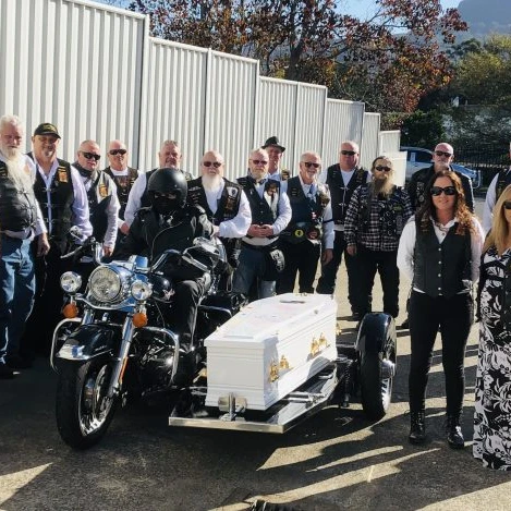 A group of people, many in biker attire, stand beside a motorbike pulling a white coffin on a trailer—a unique motorbike funeral procession, reflecting a personalised funeral service on a sunny day with white fences and trees in the background. by H Parsons Funeral Directors