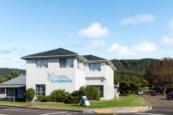 A white two-storey building with "H. Parsons Funeral Directors" signage on the side, located on a sunny street corner with green trees and hills in the background. by H Parsons Funeral Directors