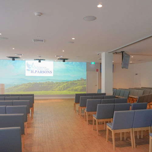 Modern funeral chapel with rows of blue-cushioned chairs facing a screen displaying “Funeral Directors H. Parsons” and an image of a green landscape. The room has wooden flooring and white walls. by H Parsons Funeral Directors