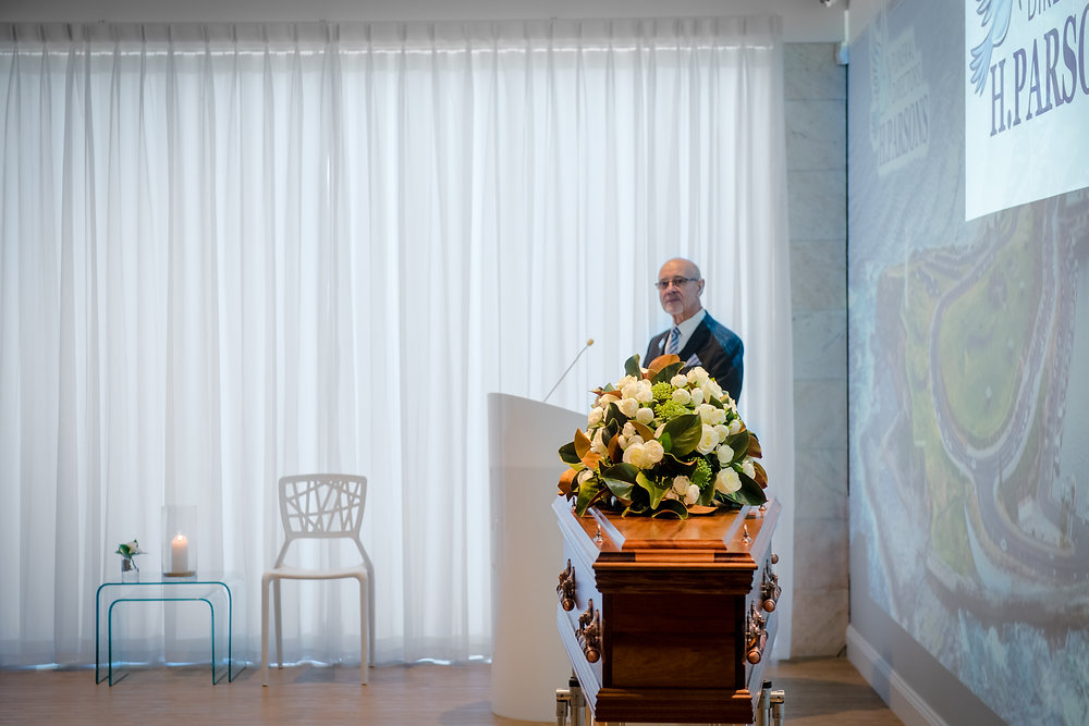 A man speaks at a lectern in a bright room with sheer curtains. In front of him is a coffin with flowers—reminding attendees not only of love lost, but also prompting reflection on how much a funeral is and the average funeral cost today. by H Parsons Funeral Directors